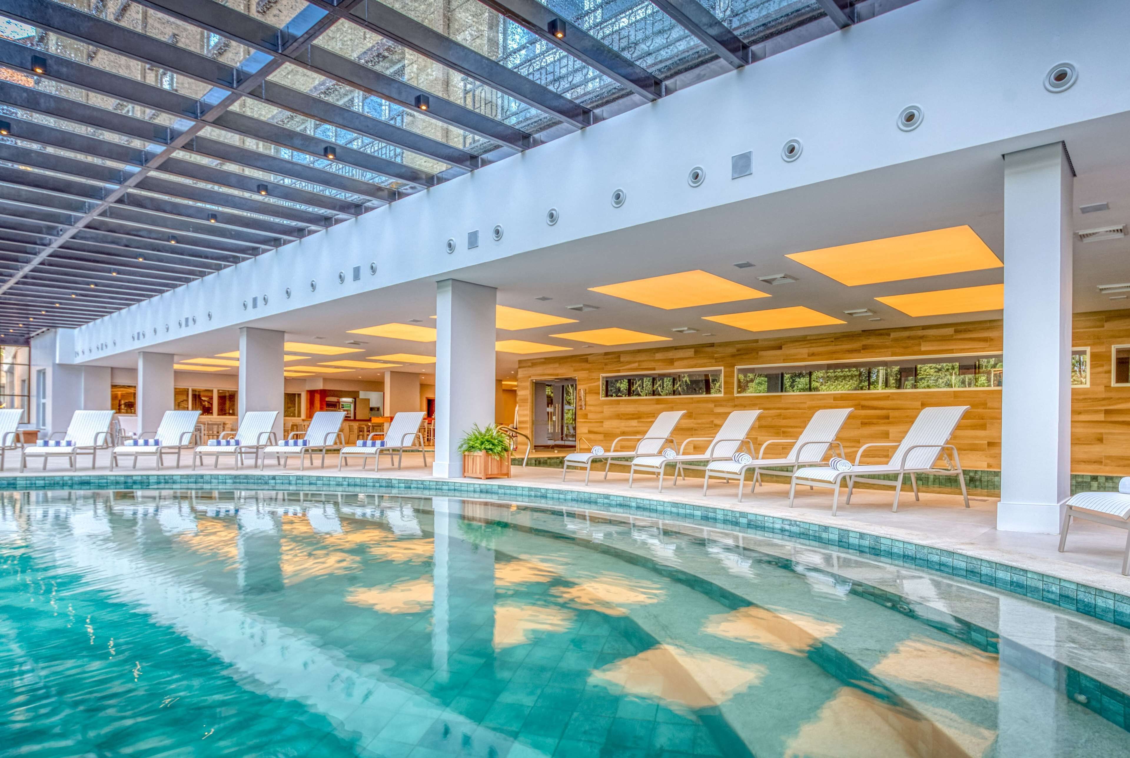 A modern indoor swimming pool with clear blue water, surrounded by white lounge chairs and a wooden wall.