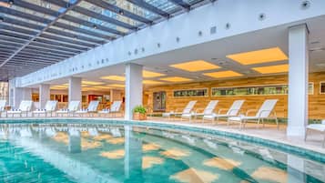 A modern indoor swimming pool with clear blue water, surrounded by white lounge chairs and a wooden wall.