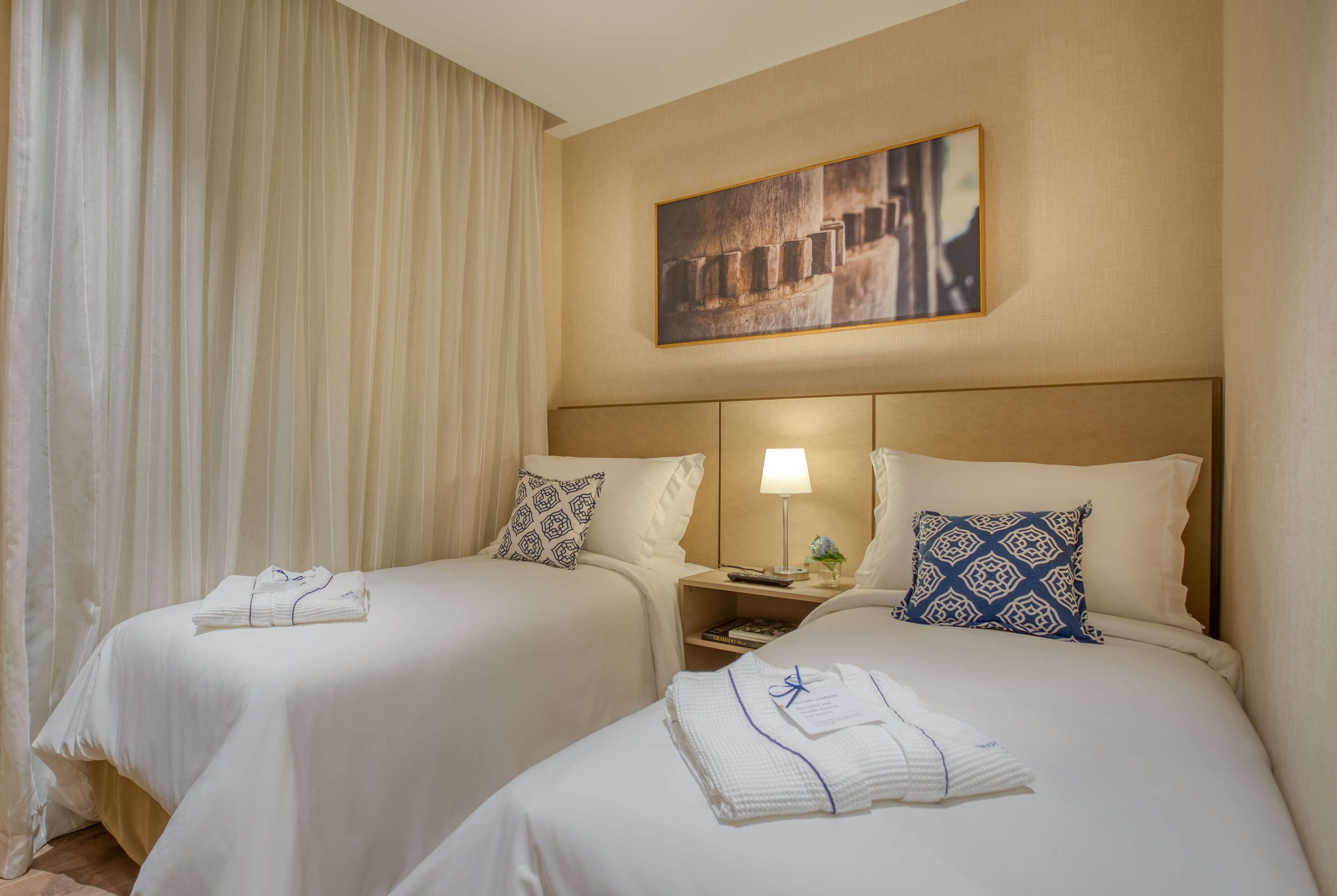 Une chambre d’hôtel avec deux lits, chacun étant équipé d’un oreiller et d’une serviette pliée. Il y a une table de chevet avec une lampe et une petite plante. Un tableau est accroché au mur au-dessus des lits.