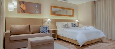 Habitación de hotel con sofá, reposapiés y cama con ropa de cama blanca. Se observa un espejo grande sobre la cama y un reloj en la pared.