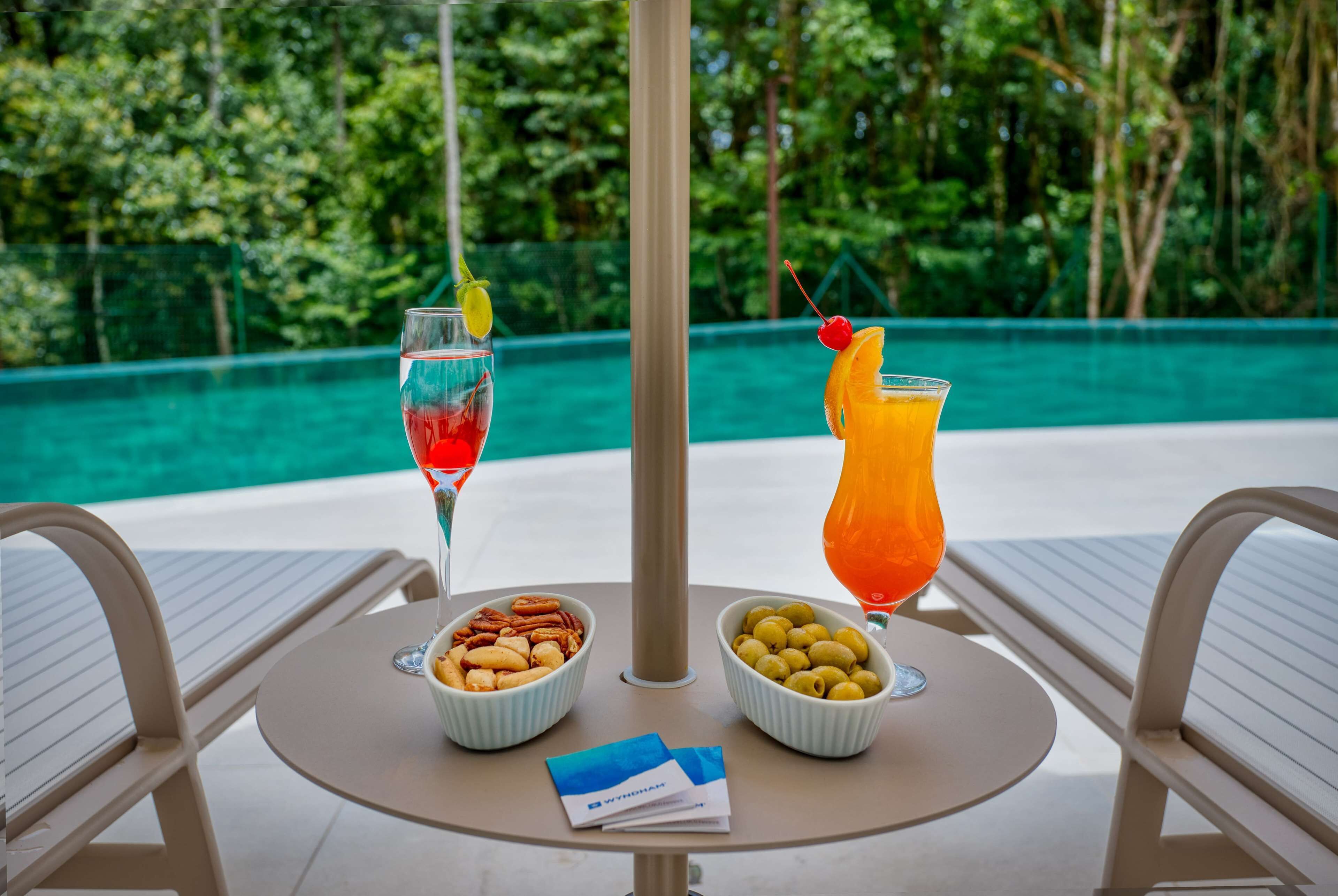 Une table avec deux bols de fruits secs et un verre d’une boisson rouge, ainsi qu’un autre verre d’une boisson orange, décoré d’une cerise, le tout placé devant une vue sur la piscine.