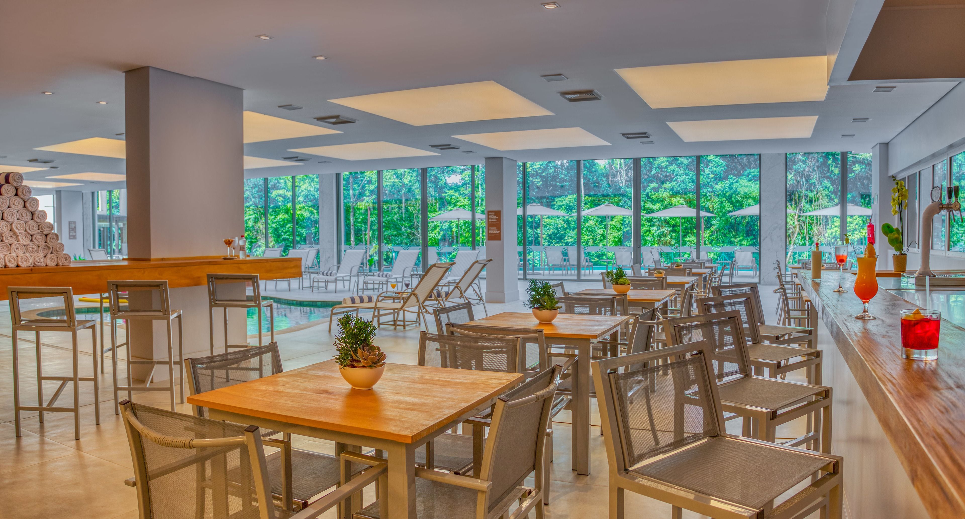 A spacious indoor area with multiple tables and chairs, large windows offering a view of greenery outside, and a bar area with a counter and seating.