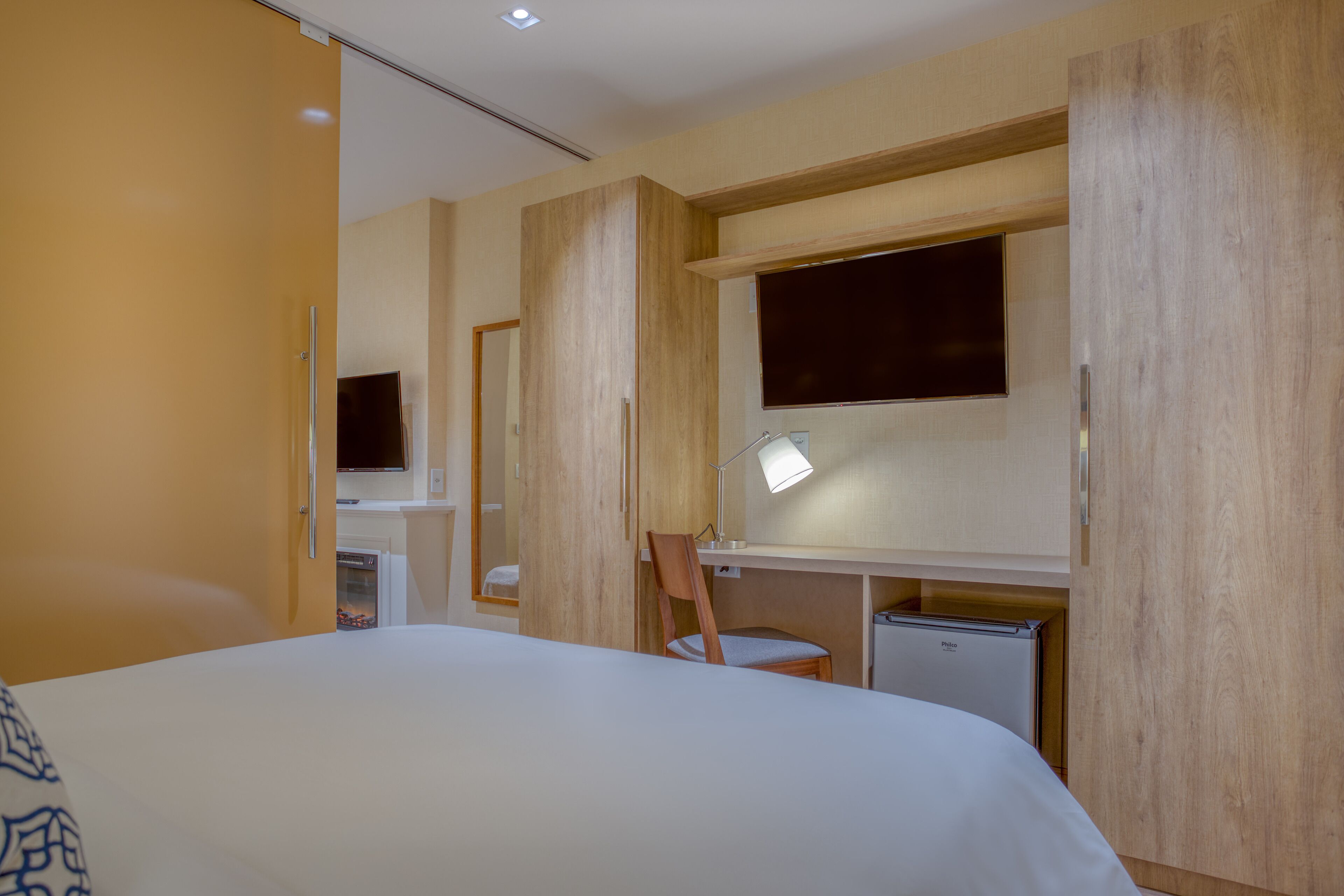 A hotel room with a bed, a desk with a chair, a television, and a small refrigerator.