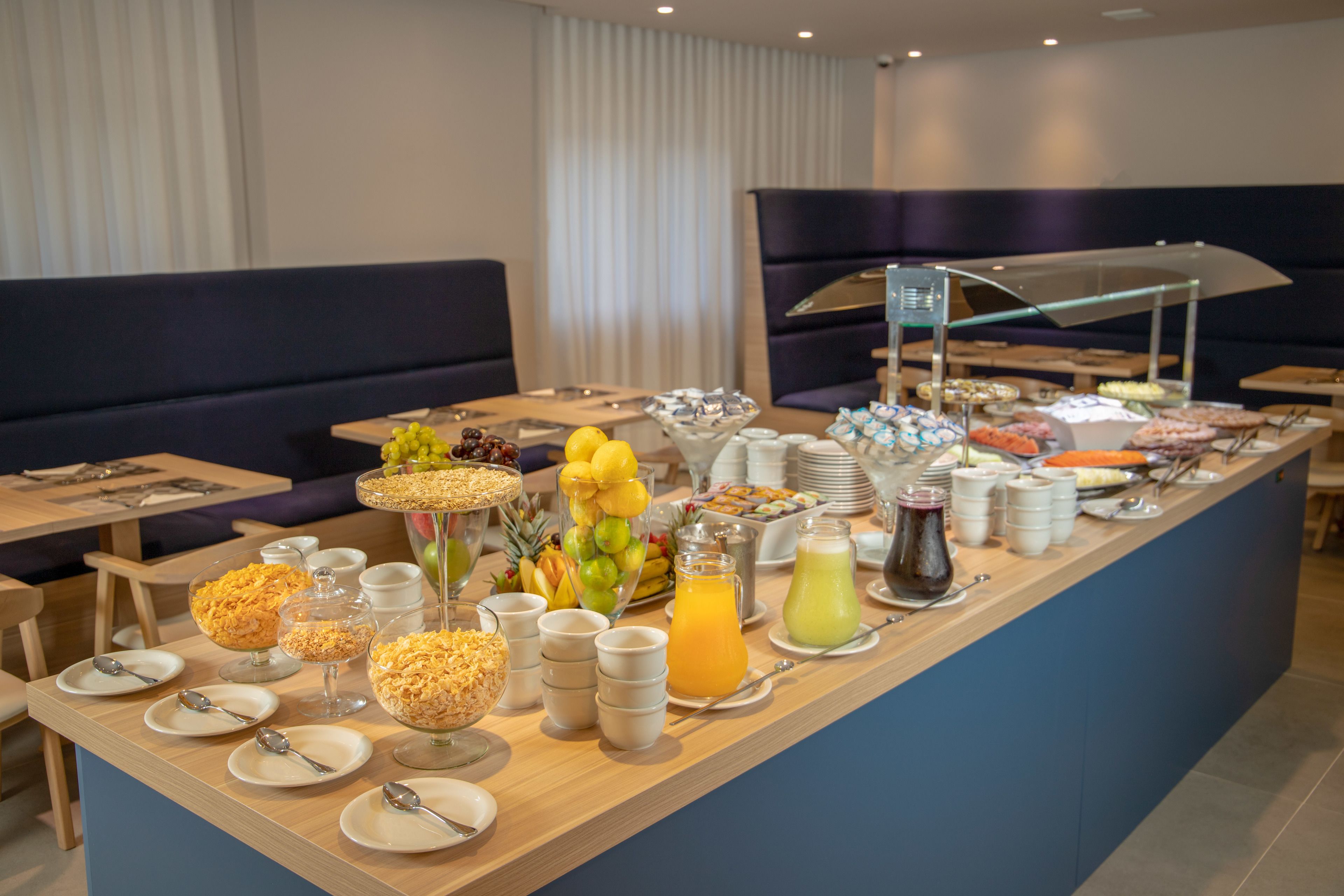 Disposição de buffet com diversos alimentos e bebidas, incluindo frutas, cereais e um dispensador de sucos.