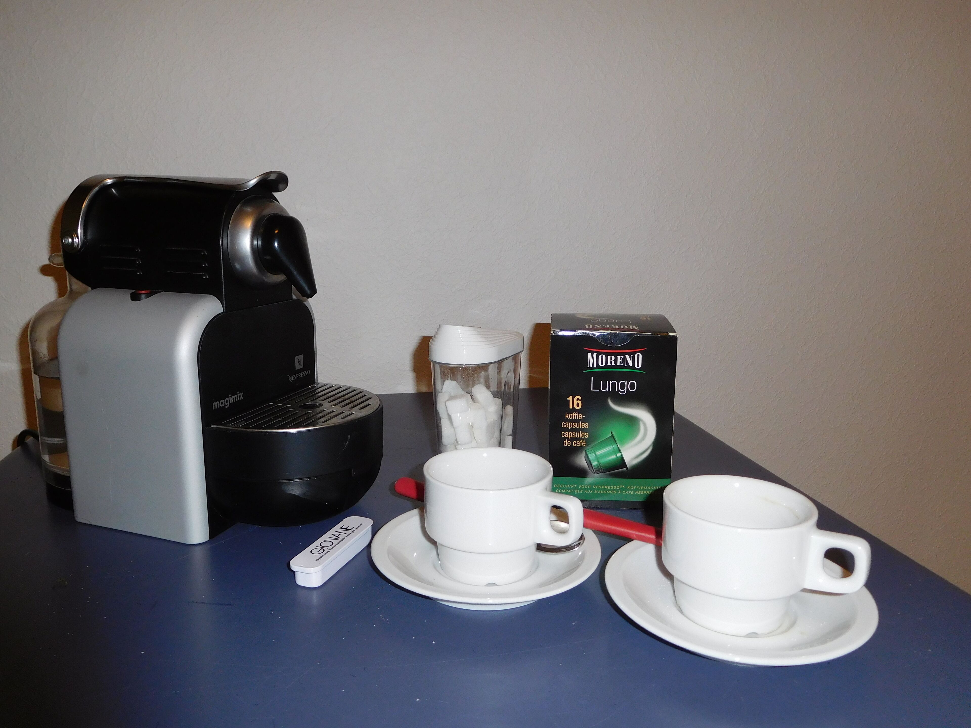 Une machine à café, deux tasses, une boîte de capsules de café et un sucrier sur une table.