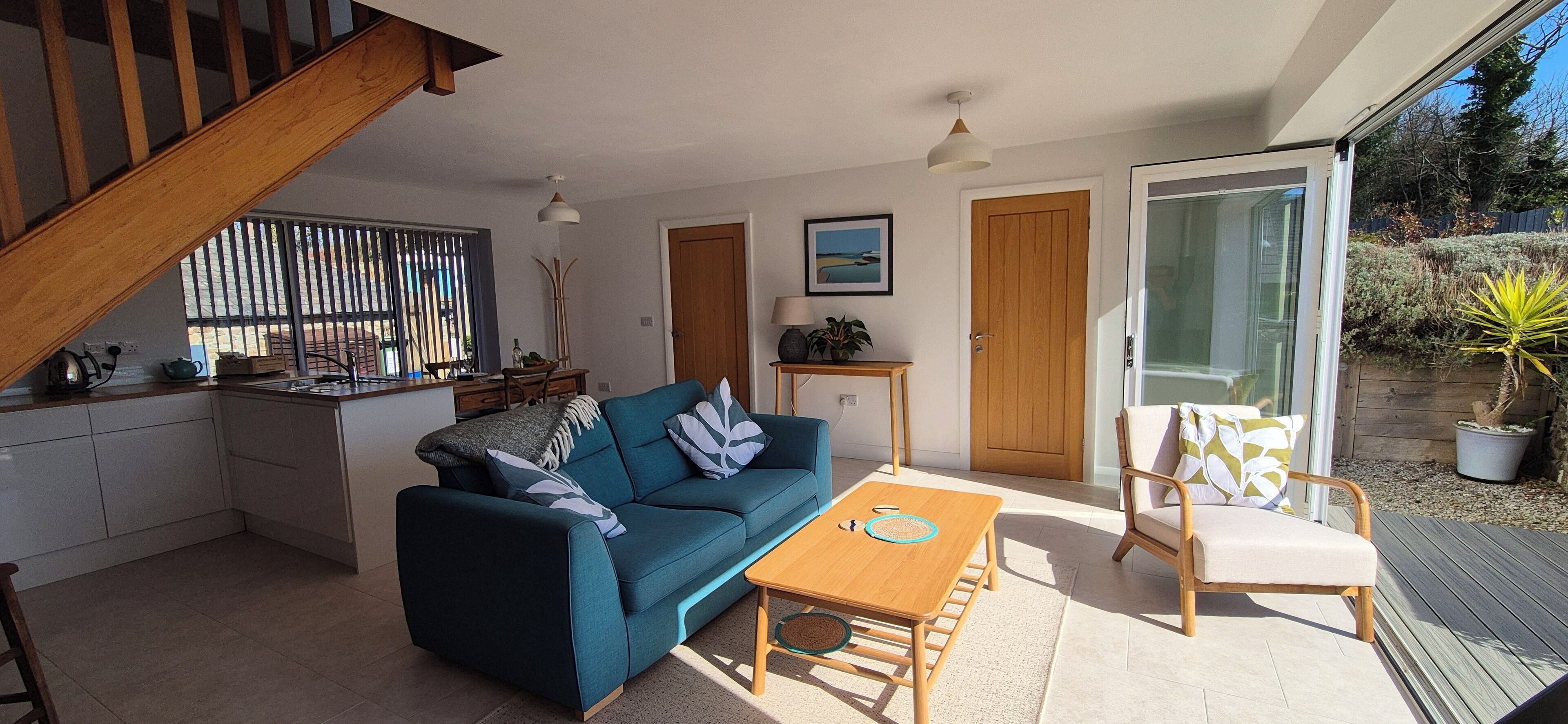 A modern living room with a sofa, coffee table, and a chair. There is a kitchen area with a dining table and chairs. The room has a large sliding glass door leading to an outdoor area with plants.