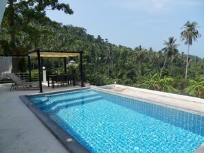 Ein Swimmingpool mit Blick auf eine üppige tropische Landschaft, eine überdachte Sitzgelegenheit und ein Tisch mit Stühlen.