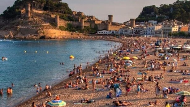 A crowded beach with people sunbathing and swimming, surrounded by historic buildings and a castle on a hill.