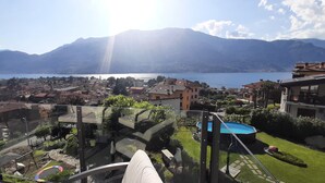 A balcony with a glass railing overlooking a residential area, a swimming pool, and a lake with mountains in the background. - Villa Mari, detached villa with magnificent garden view (Dervio)