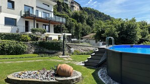 A modern outdoor pool area with a stone fire pit and a wooden deck leading to a building. - Villa Mari, detached villa with magnificent garden view (Dervio)