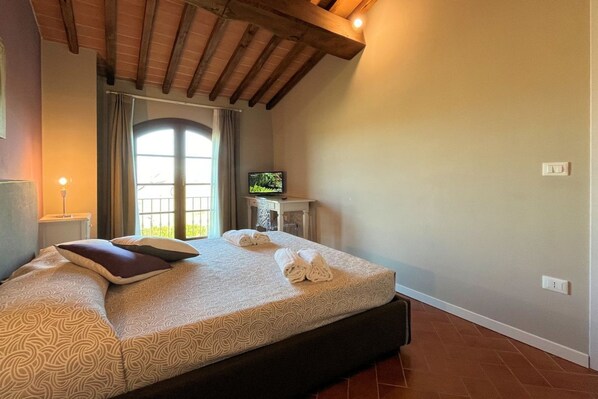Un dormitorio con cama, televisión, ventana y techo con vigas a la vista.