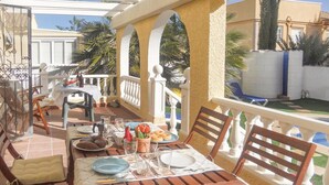 A sunlit outdoor dining area with a table set for two, surrounded by wooden chairs and a white railing.