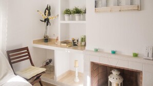 A cozy room with a wooden chair, a white bed, a fireplace, and shelves with plants and candles.