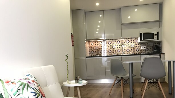 Ein kompakter Wohnbereich mit weißem Sofa, einem kleinen runden Tisch und einer Küchenzeile, die eine Mikrowelle und eine gemusterte Spritzschutzwand bietet.