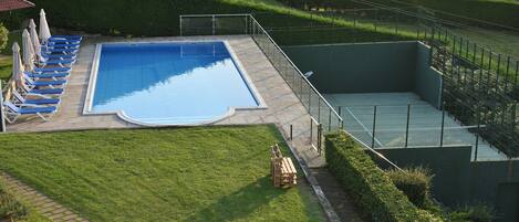 En swimmingpool med solstolar och en träbänk, omgiven av en välskött gräsmatta och ett hus i bakgrunden.