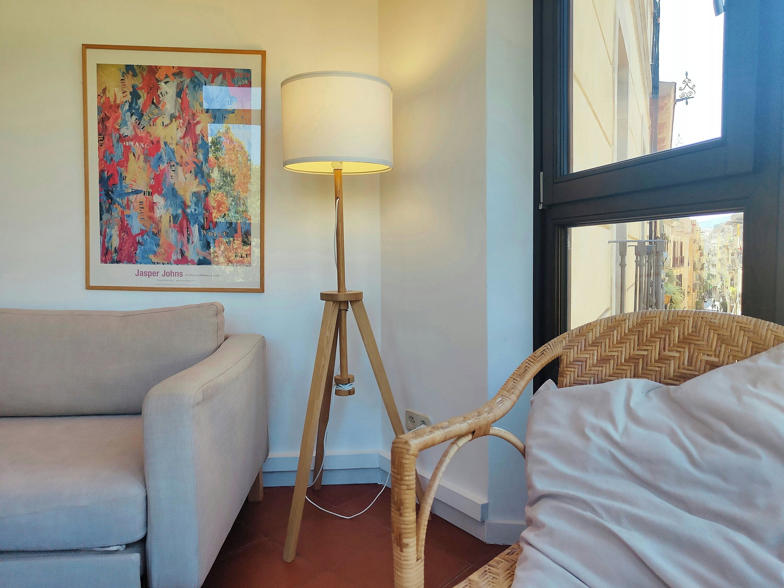 Una habitación con sofá, lámpara de pie, sillón de mimbre y ventana con vistas a edificios.