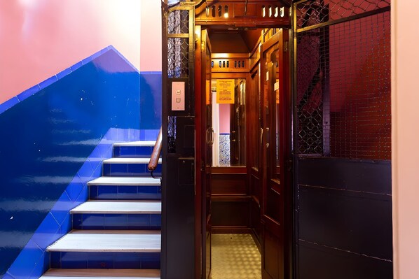 A narrow, wooden stairway leading to an elevator, with a blue-tiled wall on one side and a red-tiled wall on the other.