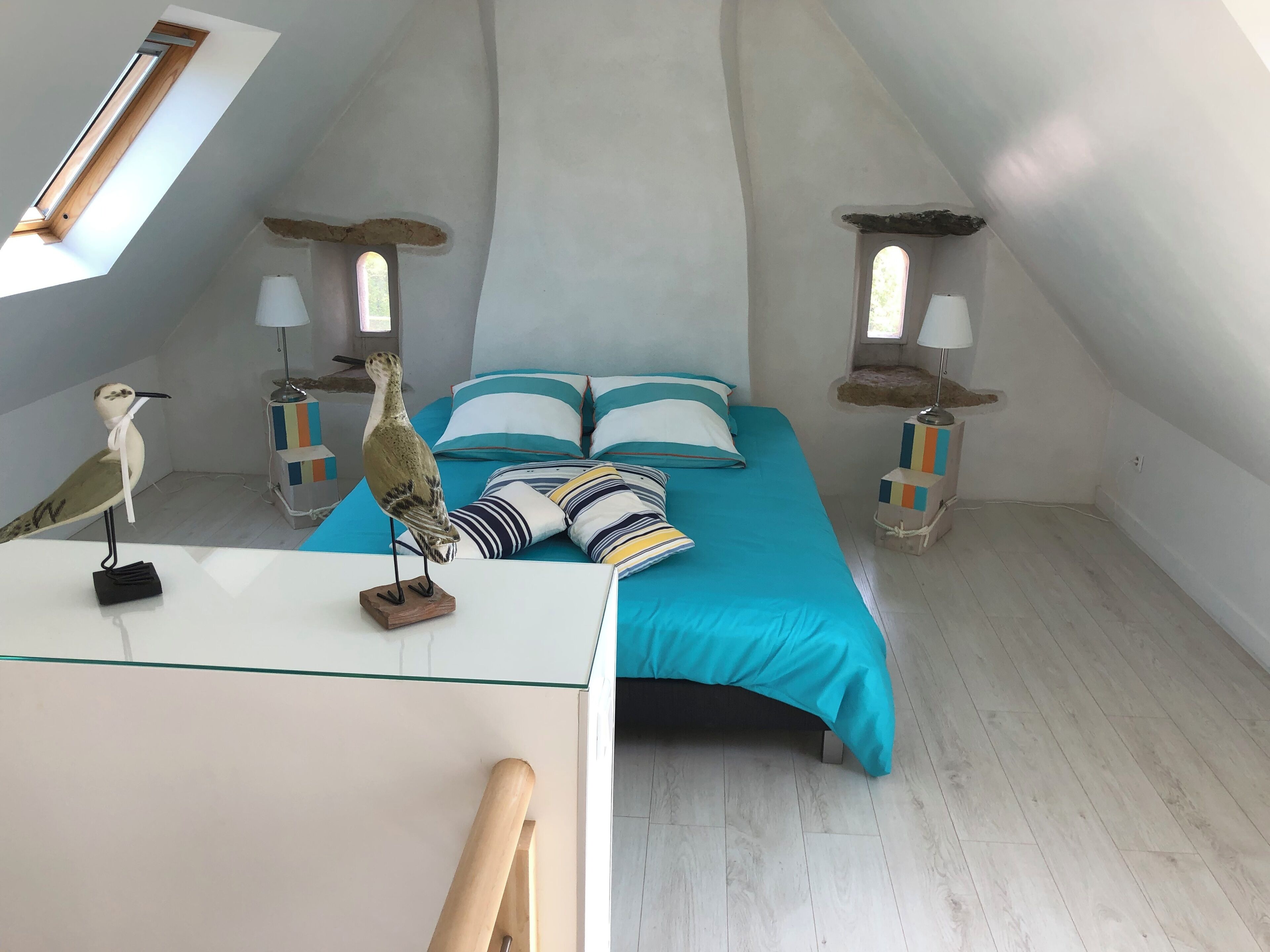 A bedroom with a sloped ceiling, a bed with a blue bedspread, striped pillows, and two bedside lamps. A wooden bird sculpture is on the bedside table.
