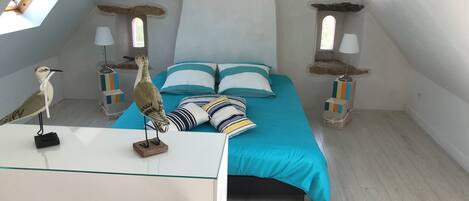 A bedroom with a sloped ceiling, a bed with a blue bedspread, striped pillows, and two bedside lamps. A wooden bird sculpture is on the bedside table.