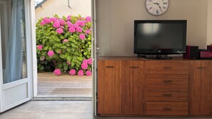 An open sliding door leading to a balcony with pink flowers, a wooden cabinet with a TV, and a wall clock.