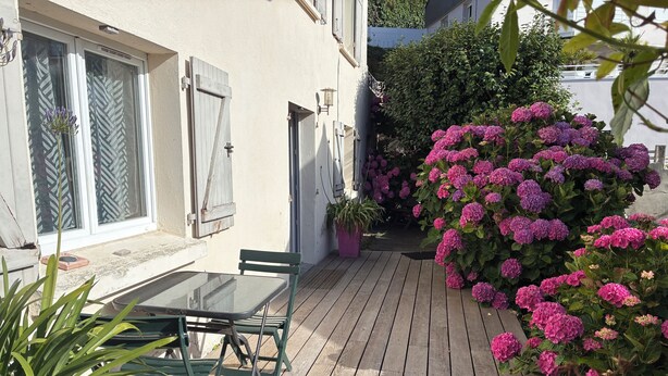 A wooden deck with a table and chairs, a potted plant, and a bush of pink flowers.