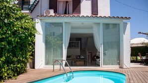 A swimming pool with a wooden deck, a covered outdoor seating area, and a building with large windows and a balcony.