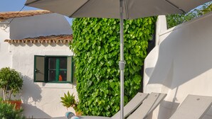 A patio with white lounge chairs and a large umbrella, surrounded by greenery and a white building with a tiled roof.