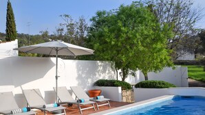 A swimming pool with lounge chairs and an umbrella, surrounded by greenery and a white wall.