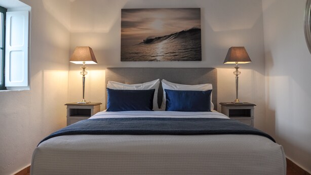 A neatly made bed with a blue and white duvet, two bedside lamps, and a painting of a seascape above the bed.