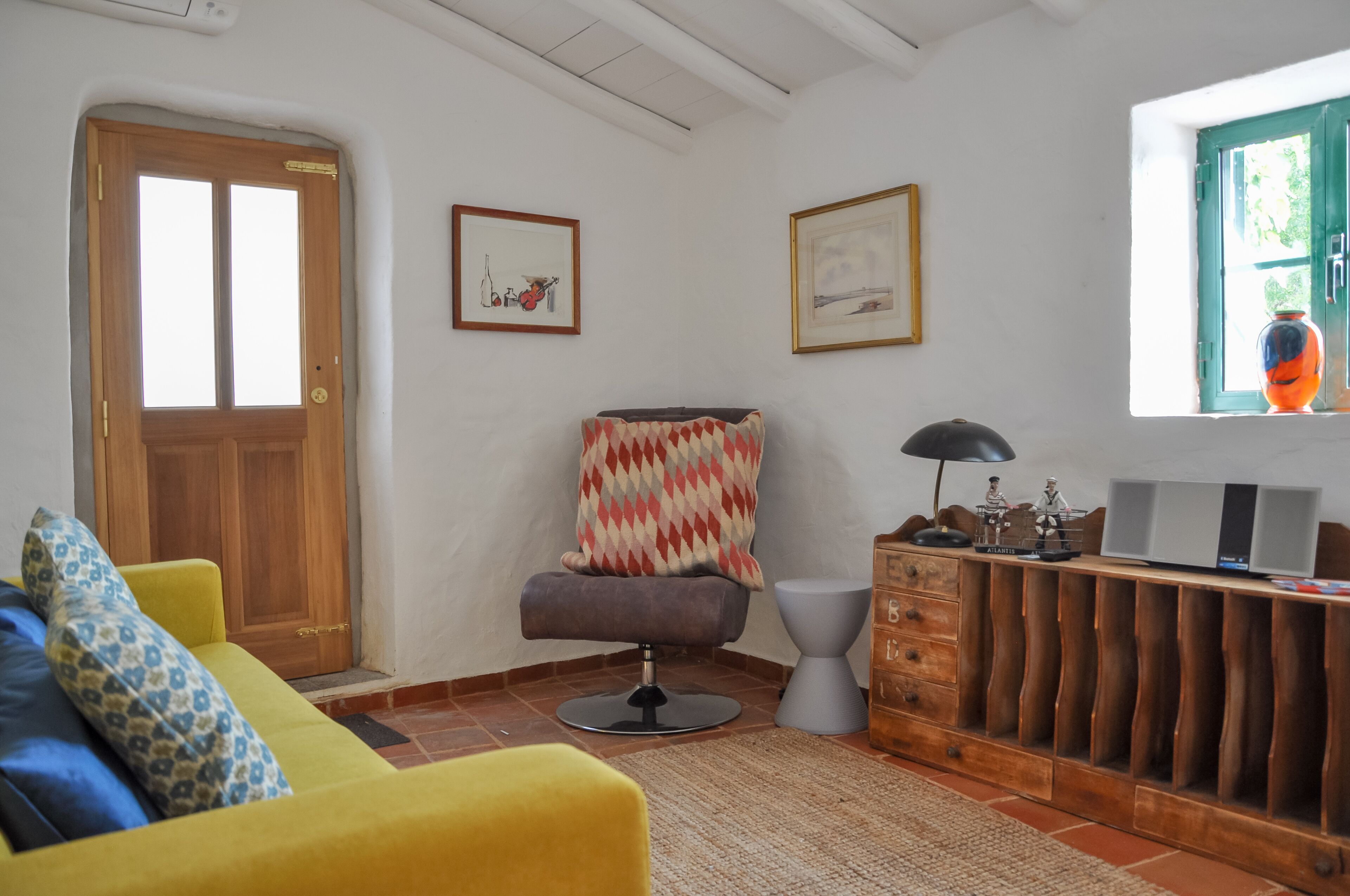 Ein Zimmer mit einer Holztür, einem gelben Sofa, einem grauen Sessel, einem Holzschrank und einem Fenster mit einer Vase.