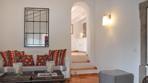 A living room with a sofa, ottoman, and coffee table, a staircase leading to a kitchen, and a wall-mounted light fixture.