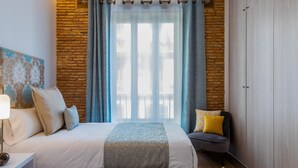 A bedroom with a brick wall, a large window with curtains, two beds, a bedside table with a lamp, and a chair with cushions. - A la luna de Valencia, 4-6 pax, 2 Bedrooms & 2 Bathrooms in downtown Valencia (Valencia)