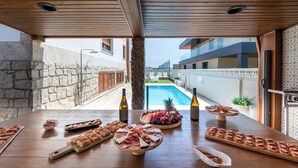 Une table en bois, sur laquelle sont disposés divers aliments et des bouteilles, donnant sur une piscine et des bâtiments modernes.
