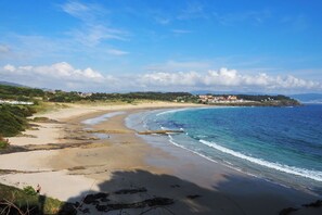 A coastal landscape with a sandy beach, a clear blue sky, and a distant view of buildings on a hill. - VILLA MONTALVO BEACH SANXENXO-HEATED SWIMMING POOL (Sanjenjo-Pontevedra)