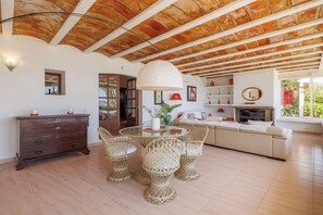 A room with a wooden ceiling, a round table with wicker chairs, a sofa, a TV, and a small cabinet.