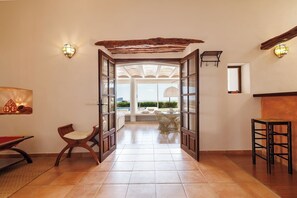 An open doorway leading to a sunlit room with a view of the outdoors, a wooden bench, a small table, and wall-mounted lights.