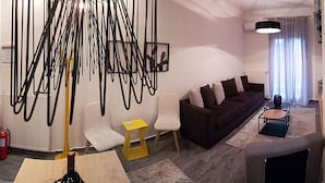 A modern living room with a dark brown sofa, white chairs, and a yellow side table. There is a black pendant light and a fire extinguisher on the wall. - Alpinia Deluxe Apt Must (Thessaloniki)