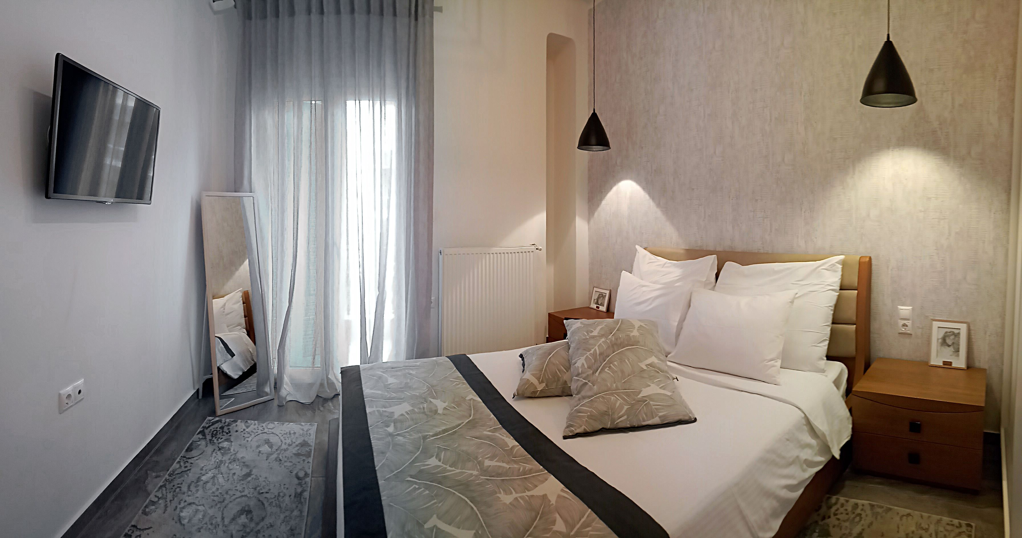 A modern hotel room with a large bed, a bedside table, a nightstand, a wall-mounted TV, and a window with sheer curtains.