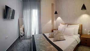 A modern hotel room with a large bed, a bedside table, a nightstand, a wall-mounted TV, and a window with sheer curtains. - Alpinia Deluxe Apt Must (Thessaloniki)