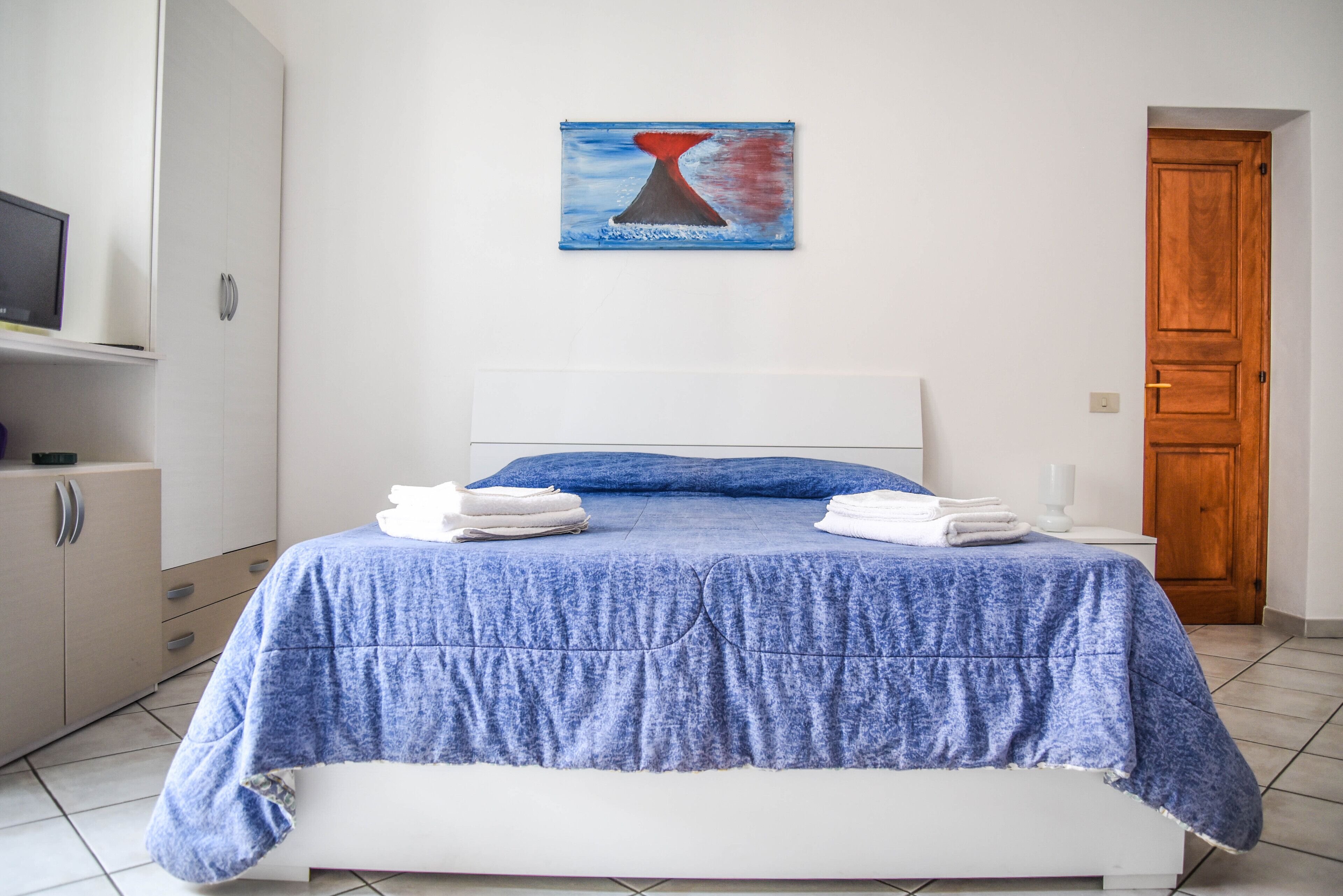 A bedroom with a bed, a painting on the wall, a wooden door, and a white wardrobe.