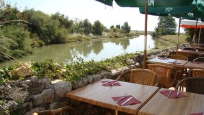 An outdoor dining area with tables and wicker chairs, overlooking a calm river and surrounded by greenery. - Villa 3 by Avecoeur (Saint-Augustin)