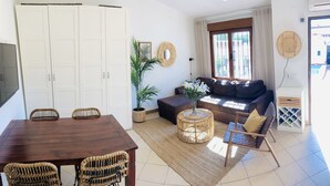 Una sala de estar con sofá, mesa de centro, mesa de comedor con sillas y un ventanal con cortinas.