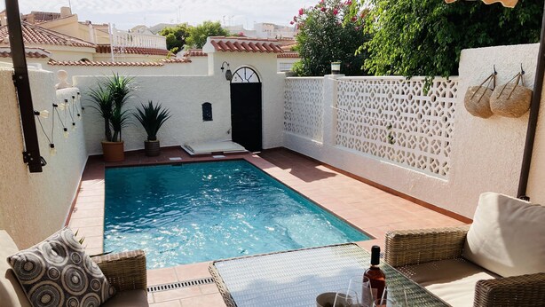 Un área de piscina con mesa de vidrio, mobiliario de mimbre y una botella de vino.
