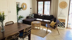 Una sala de estar con un sofá de cuero marrón, una mesa de comedor con sillas de mimbre, un ventanal con cortinas y un ventilador de pared.