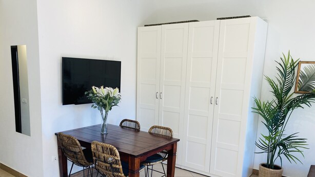 Un comedor con una mesa de madera, sillas de mimbre, un jarrón con flores, un televisor de pantalla plana, armarios blancos y una planta en maceta.