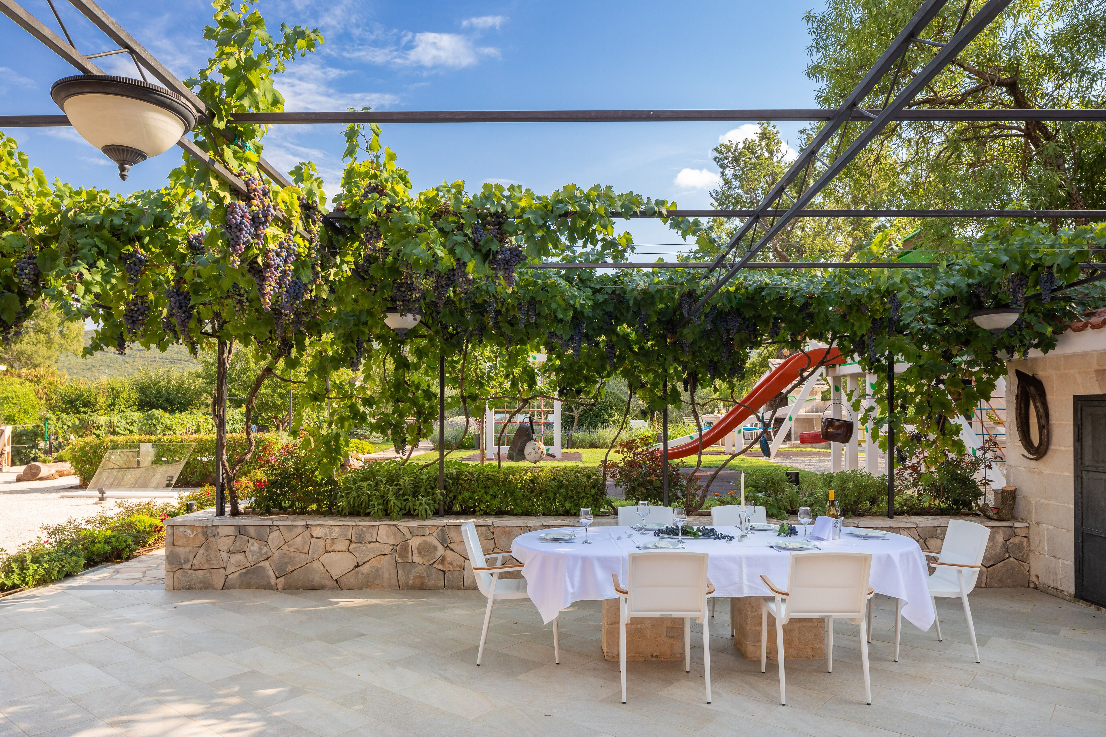 An outdoor dining area with a table set for four, surrounded by stone walls and a pergola with grapevines.