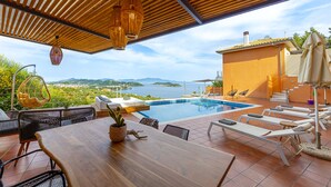 A poolside area with a wooden table, lounge chairs, and a view of the sea. - IO.DELUXE 2ΒR, 2 BTHR.VILLA WITH PRIVATE POOL  AND STUNNING SEA VIEWS  (SKIATHOS)