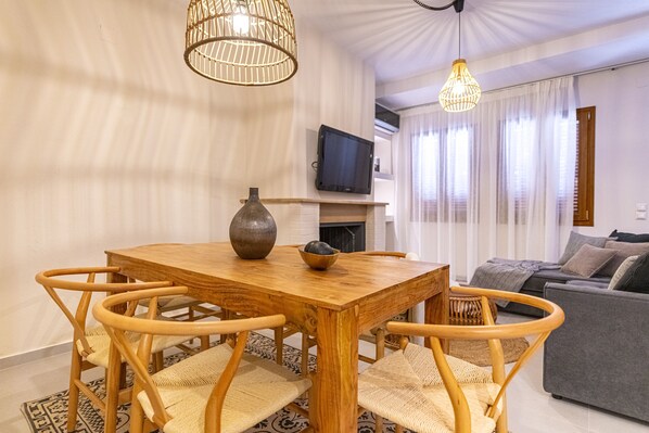 Une table de salle à manger en bois, avec des chaises, un vase et un saladier. On aperçoit un téléviseur sur une étagère au-dessus de la cheminée. Un canapé et des rideaux sont visibles à l’arrière-plan.