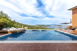 Una piscina con vistas al mar y colinas, un área de descanso con un sofá y cojines, y una silla.