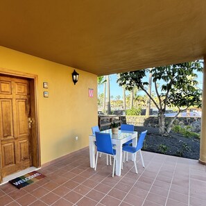 Une terrasse couverte avec une porte en bois, un paillasson, une table avec des chaises bleues, et une vue sur des palmiers et un mur de pierre.